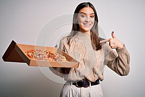 Young beautiful brunette woman holding box with italian pizza over white background with surprise face pointing finger to himself