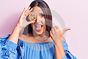 Young beautiful brunette woman holding cookie pointing thumb up to the side smiling happy with open mouth