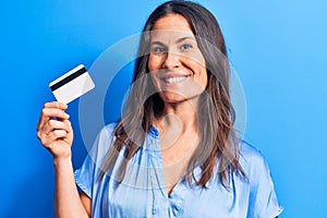 Young beautiful brunette woman holding credit card over isolated blue background looking positive and happy standing and smiling