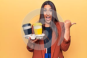 Young beautiful brunette woman holding cup of takeaway coffee pointing thumb up to the side smiling happy with open mouth