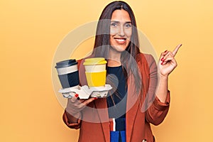 Young beautiful brunette woman holding cup of takeaway coffee smiling happy pointing with hand and finger to the side