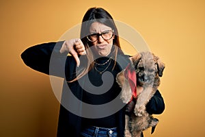 Young beautiful brunette woman holding cute puppy pet over isolated yellow background with angry face, negative sign showing
