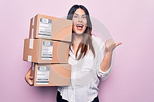 Young beautiful brunette woman holding deliver packages standing over pink background pointing thumb up to the side smiling happy