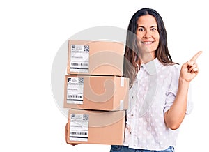 Young beautiful brunette woman holding delivery cardboard package smiling happy pointing with hand and finger to the side
