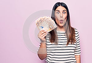 Young beautiful brunette woman holding euros banknotes over isolated pink background scared and amazed with open mouth for