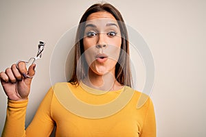 Young beautiful brunette woman holding eyelash curler over isolated white background scared in shock with a surprise face, afraid