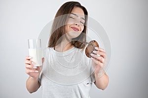 Young beautiful brunette woman holding glass of milk in one hand and tasty chocolate cookie in another looking satisfied healthy