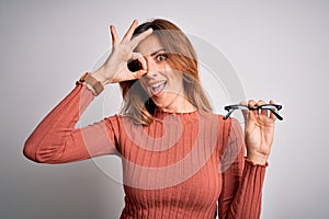 Young beautiful brunette woman holding glasses over isolated white background with happy face smiling doing ok sign with hand on