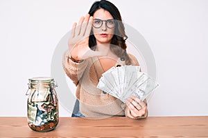 Young beautiful brunette woman holding jar with savings holding dollars with open hand doing stop sign with serious and confident