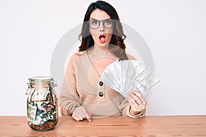 Young beautiful brunette woman holding jar with savings holding dollars scared and amazed with open mouth for surprise, disbelief