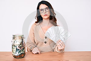 Young beautiful brunette woman holding jar with savings holding dollars thinking attitude and sober expression looking self