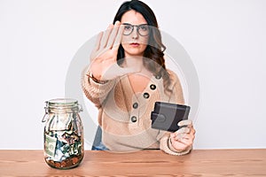 Young beautiful brunette woman holding jar with savings holding wallet with open hand doing stop sign with serious and confident