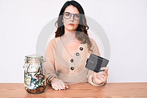 Young beautiful brunette woman holding jar with savings holding wallet thinking attitude and sober expression looking self