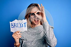 Young beautiful brunette woman holding paper with brexit message over blue background with happy face smiling doing ok sign with