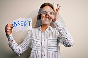 Young beautiful brunette woman holding paper with brexit message over white background with happy face smiling doing ok sign with