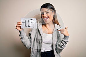 Young beautiful brunette woman holding paper with help message over white background with surprise face pointing finger to himself