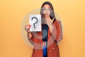 Young beautiful brunette woman holding paper with question mark covering mouth with hand, shocked and afraid for mistake