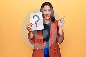 Young beautiful brunette woman holding paper with question mark smiling happy pointing with hand and finger to the side