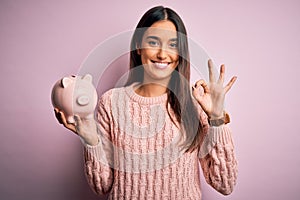 Young beautiful brunette woman holding piggy bank saving money for retirement doing ok sign with fingers, excellent symbol