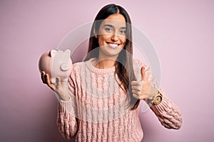 Young beautiful brunette woman holding piggy bank saving money for retirement happy with big smile doing ok sign, thumb up with