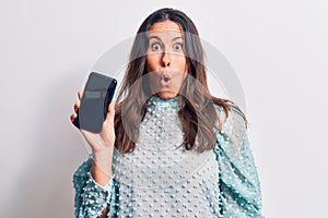 Young beautiful brunette woman holding smartphone showing screen over white background scared and amazed with open mouth for