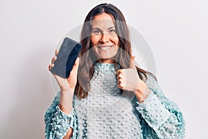 Young beautiful brunette woman holding smartphone showing screen over white background smiling happy and positive, thumb up doing