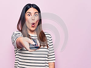 Young beautiful brunette woman holding television remote control over pink background scared and amazed with open mouth for