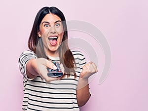 Young beautiful brunette woman holding television remote control over pink background screaming proud, celebrating victory and