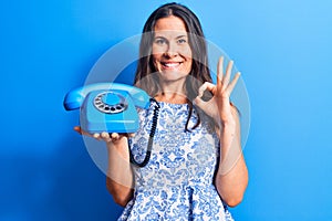 Young beautiful brunette woman holding vintage telephone over isolated blue background doing ok sign with fingers, smiling