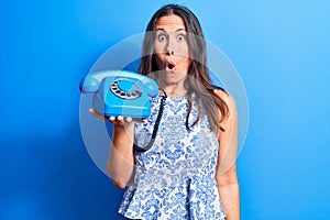 Young beautiful brunette woman holding vintage telephone over isolated blue background scared and amazed with open mouth for