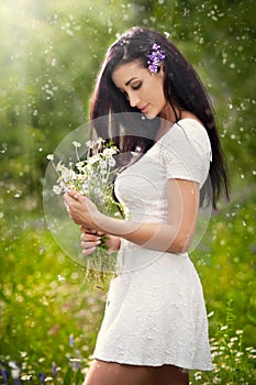 Young beautiful brunette woman holding a wild flowers bouquet in a sunny day. Portrait of attractive long hair female in white