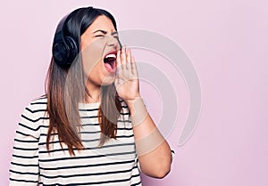 Young beautiful brunette woman listening to music using headphones over pink background shouting and screaming loud to side with