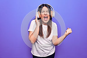Young beautiful brunette woman listening to music using headphones over purple background screaming proud, celebrating victory and