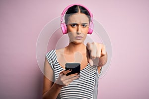 Young beautiful brunette woman listening to music using headphones and smartphone pointing with finger to the camera and to you,