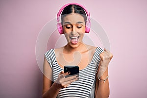 Young beautiful brunette woman listening to music using headphones and smartphone screaming proud and celebrating victory and