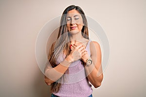 Young beautiful brunette woman with long hair standing over isolated background smiling with hands on chest with closed eyes and