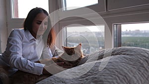 Young beautiful brunette woman lying on the bed by the window stroking a ginger Maine Coon cat that lies next to