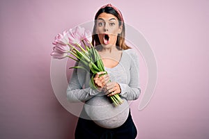 Young beautiful brunette woman pregnant expecting baby holding bouquet of tulips scared in shock with a surprise face, afraid and