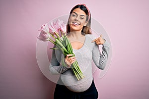 Young beautiful brunette woman pregnant expecting baby holding bouquet of tulips with surprise face pointing finger to himself