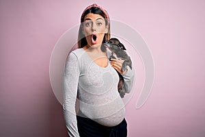 Young beautiful brunette woman pregnant expecting baby holding chihuahua puppy scared in shock with a surprise face, afraid and