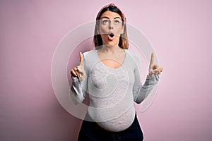 Young beautiful brunette woman pregnant expecting baby over isolated pink background amazed and surprised looking up and pointing