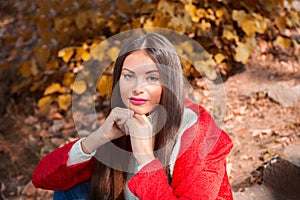 Young beautiful brunette woman with red lips and long straight hair wearing red jacket sitting outdoor in autumn park.