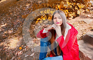 Young beautiful brunette woman with red lips and long straight hair wearing red jacket sitting outdoor in autumn park.