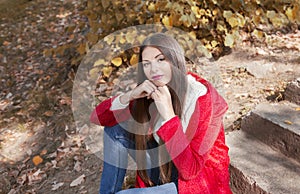 Young beautiful brunette woman with red lips and long straight hair wearing red jacket sitting outdoor in autumn park.