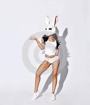 Young beautiful brunette woman in sexy white sports clothing or underwear and white rabbit mask on face standing over white wall