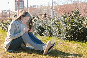 Young beautiful brunette woman sits on grass in spring park, listen music on phone and sing a song.