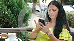 Young beautiful brunette woman sitting in an outdoor cafe, drinking coffee and using a phone