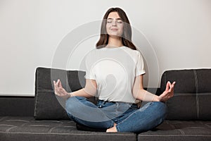 Young beautiful brunette woman sitting on the sofa in calm yoga position in her appartment looking peaceful, feels relaxed and