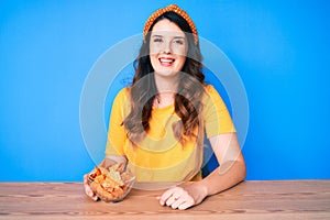 Young beautiful brunette woman sitting on the table eating nachos potato chips looking positive and happy standing and smiling