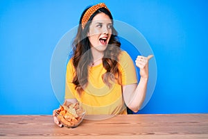 Young beautiful brunette woman sitting on the table eating nachos potato chips pointing thumb up to the side smiling happy with